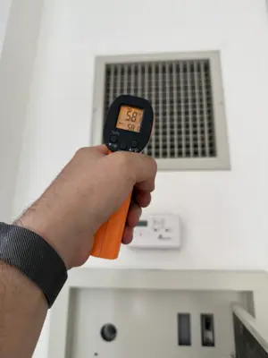 Technician using infrared thermometer to measure air vent temperature reading 58°F during HVAC inspection in NYC