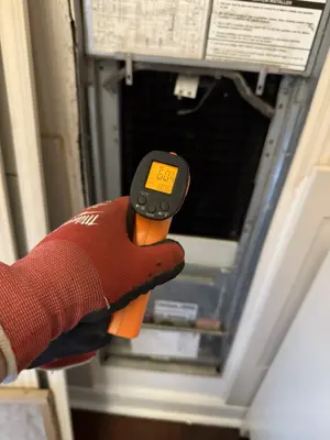 Gloved technician measuring HVAC air handler temperature with infrared thermometer reading 60°F during AC service