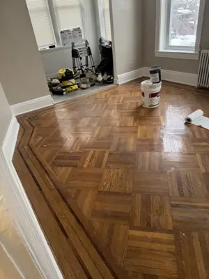 Parquet hardwood floor refinishing in progress in a NYC apartment with tools and finish bucket visible