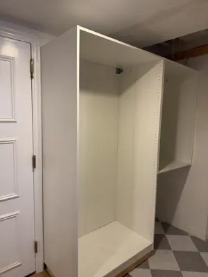 Assembled white IKEA PAX wardrobe unit installed in basement room with checkered floor in NYC