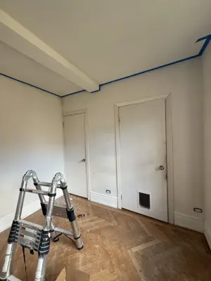 Room painting in progress with blue painter's tape on ceiling edges and ladder set up in NYC apartment