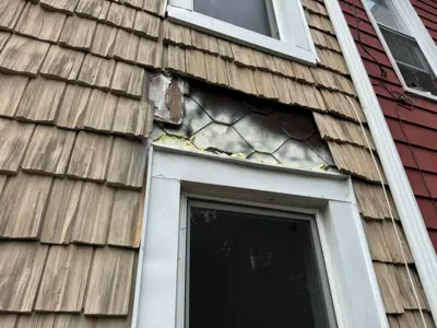 Severely damaged exterior siding above window showing exposed wire mesh, rot, and missing shingles on NYC home