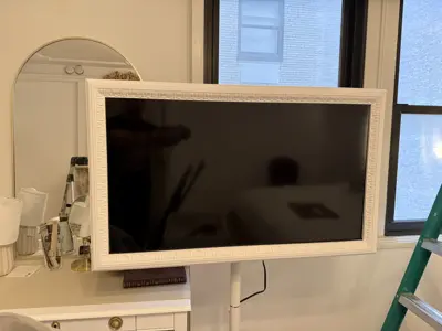 Large flat-screen TV mounted in decorative white frame on wall of a bright NYC apartment bedroom